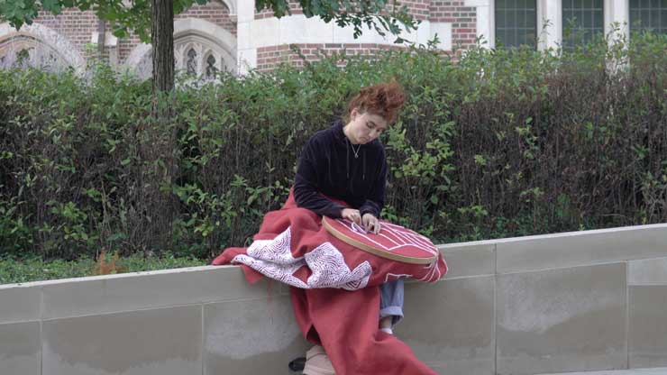 A woman doing needlepoint, sitting on a short wall.