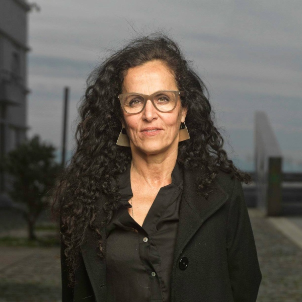Laura Bari stands in a black blouse and jacket, she has long black curly hair and wears glasses.