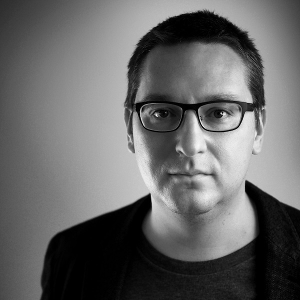 Black and white photo of a white man dressed in black and wearing dark rimmed glasses