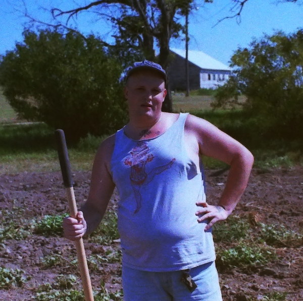 Image of artist standing in a field holding what appears to be a shovel with one hand on their hip