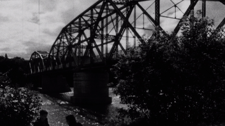 A black and white image of a steel bridge. there is a tree in the foreground, and two people sitting