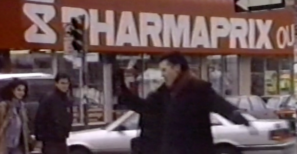 Une femme aux cheveux courts vêtue d'un manteau noir virevolte sur un trottoir enneigé près d'un magasin Pharmaprix. Un homme et une femme marchent à l'arrière-plan. 