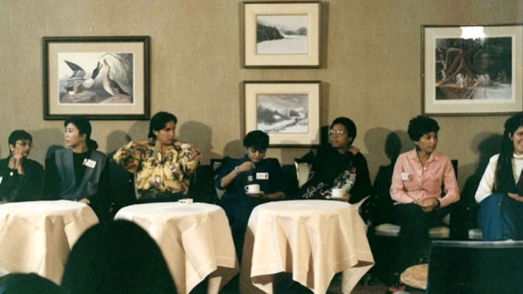 Un groupe composé de femmes de couleur discute, assises à des tables dans une pièce bien éclairée.