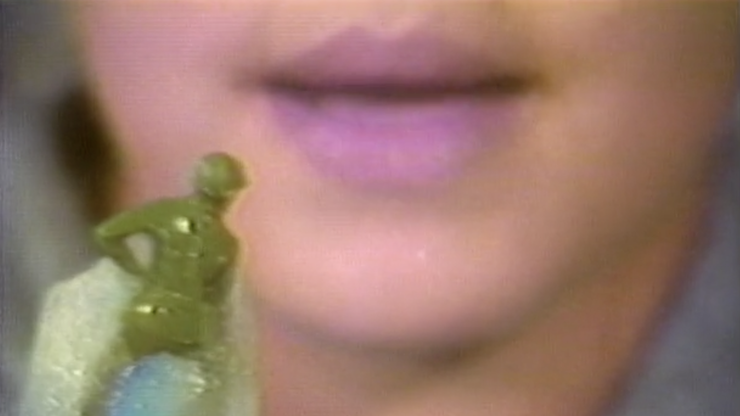 A child with pink lips holds a green soldier toy in front of their mouth