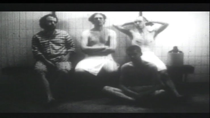 Black and white image of four men, three of which are dressed with towels tied around their waist, seemingly sitting in a bathhouse.