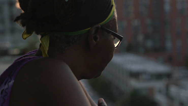 Image fixe en couleur. Une personne noire avec des lunettes et un bandeau jaune-vert regarde un paysage urbain.