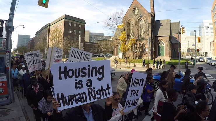 Streetview of a group of protesters holding a sign that says "Housing is a human right"