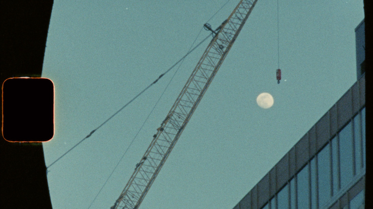 Une grue au crépuscule, la lune est encadrée entre la grue et le bâtiment dans le coin inférieur gau