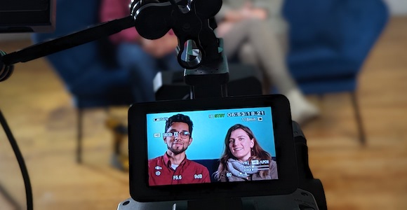 A man and a woman in a camera's viewfinder in the foreground, while they sit on a couch behind it.