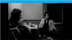 Deux personnes assises à une table, l'image est en noir et blanc, nous sommes dans les années 1970.
