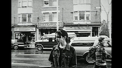 A black and white image of a young girl standing on a street with cars and storefronts behind her.