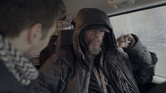 A man sits inside a vehicle wearing a jacket with the hood up and holding a cigarette.