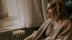 A woman in a bra and cardigan sits on a chair and looks out the window.