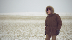 Man in brown parka, looking in the distance in front of a desolate landscape