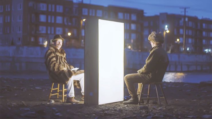 Two men sit on either side of a box emitting bright white light in the evening outside.