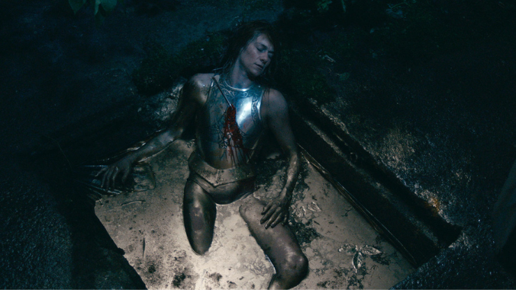 A person wearing a silver breastplate pierced by an arrow half submerged in brown liquid.