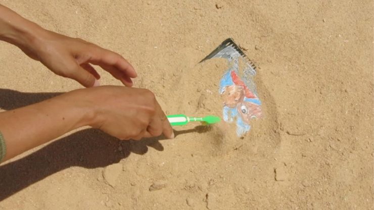 A painted object submerged in sand being cleared off by a toothbrush.