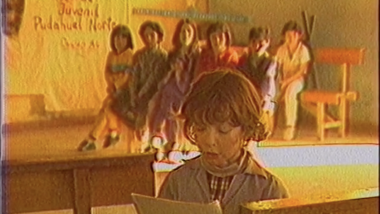 A group of young children in a classroom. One girl in the foreground is reading from pages.