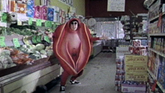 Grainy colour photo in a grocery store aisle. A person in a pink costume in the shape of a vagina.