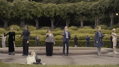 A cirlce of people in standing around a fountain each with one arm outstretched 
