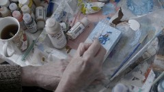 A person painting on a small eisel with different paints surrounding them