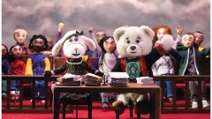 A film still of a plush lamb and a bear sitting at a desk with a cheering crowd behind them