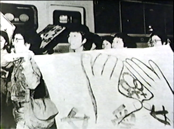 A black and white still of a group marching. A large poster of two hands holding many gender symbols is held up.