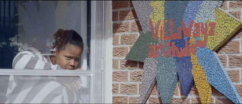 Arrêt sur image. Une personne noire regarde à travers une porte vitrée. À droite, il y a un mur de briques avec un panneau en mosaïque qui indique "Villawayz Art Studio". 